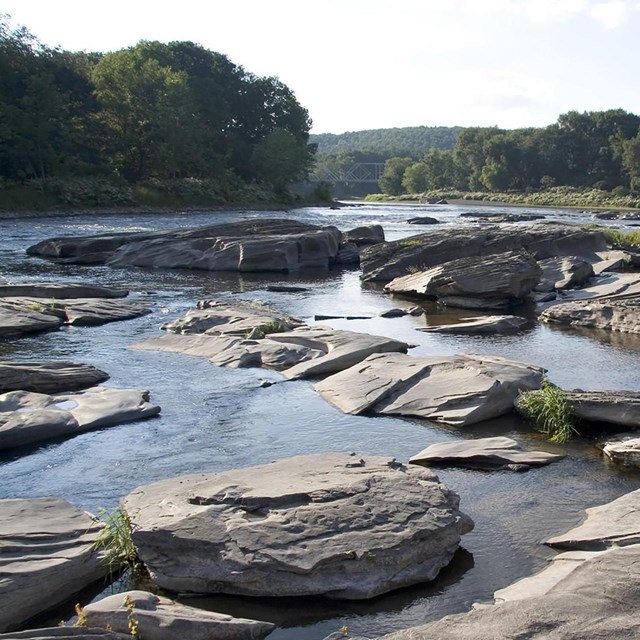 A wide shot of dark, smooth water reflecting light. Large grey stones like islands in the middle.